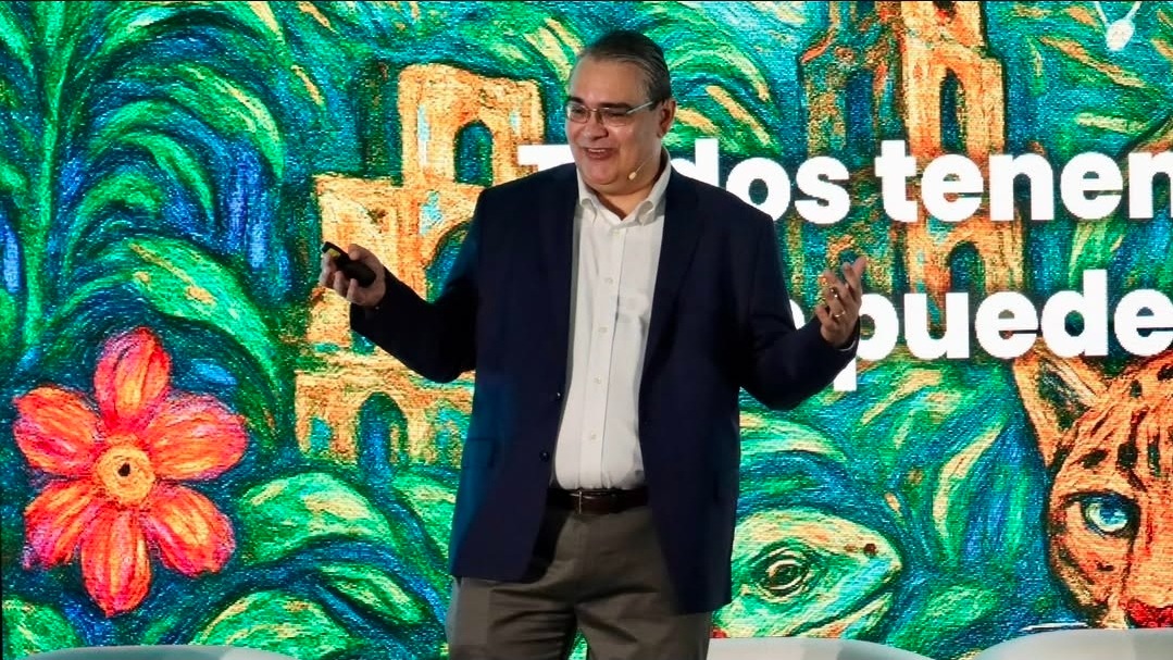 Roberto Melo hablando en un keynote con un mural artístico de fondo